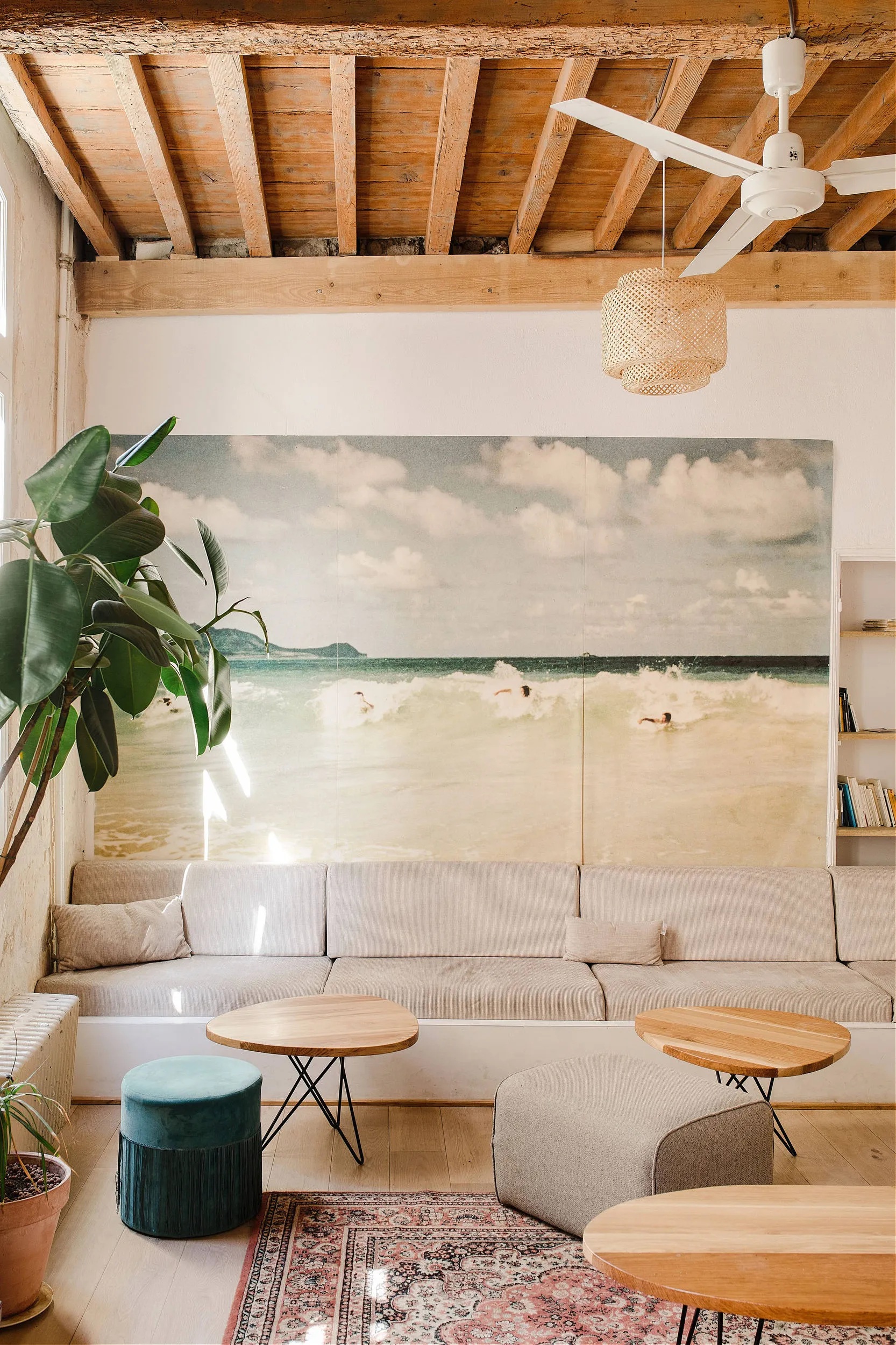 A boho-chic cafe features beige-tinted interiors with pops of color manifested across rugs, velvety stools in sea green, large-scale paintings, and plants.