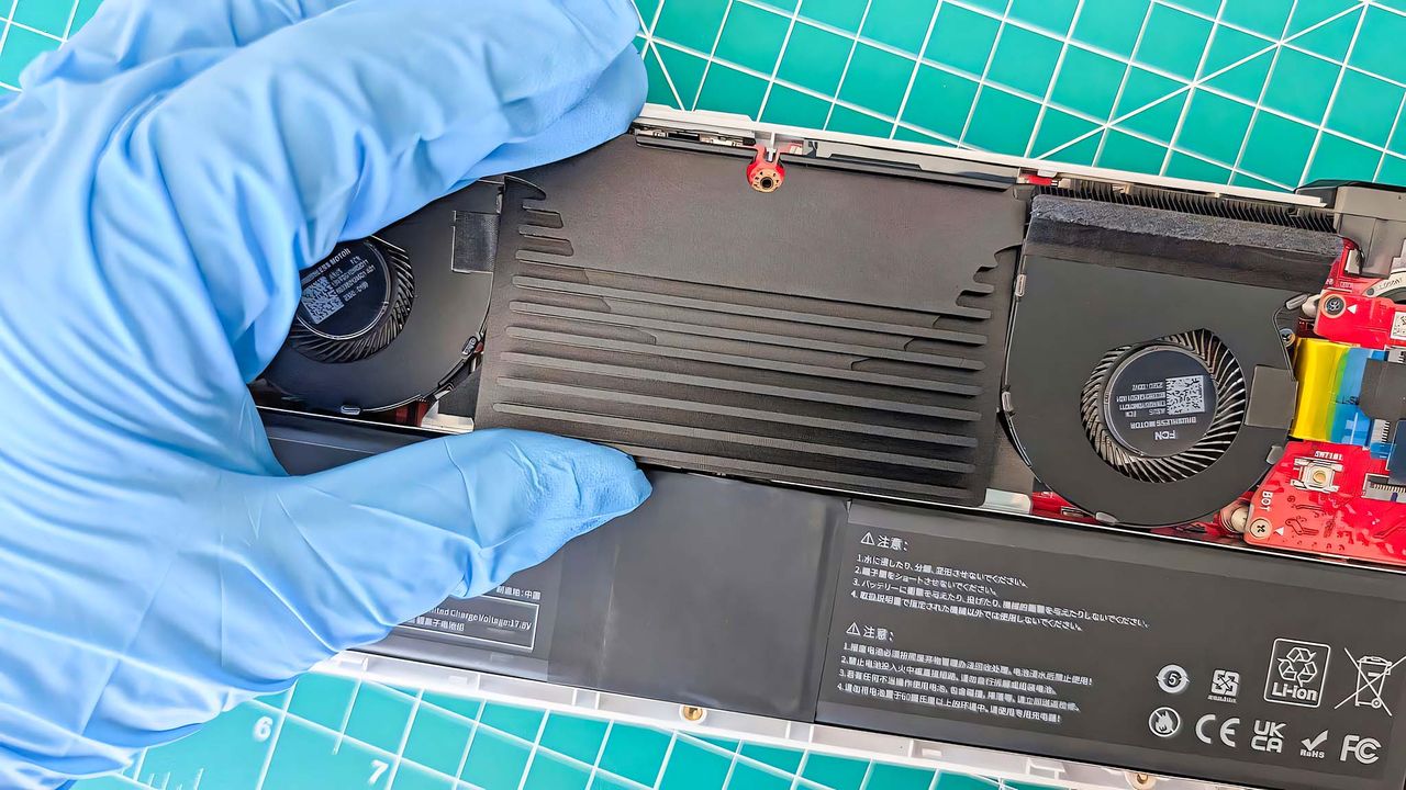 A gloved hand installing the JSAUX ROG Ally Battery Upgrade Kit's thermal plate into an open Ally.