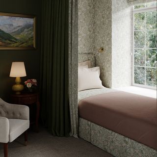 tiny guest room ideas, ditsy green leafy wallpaper, bed by window with curtain across, armchair, artwork, lamp