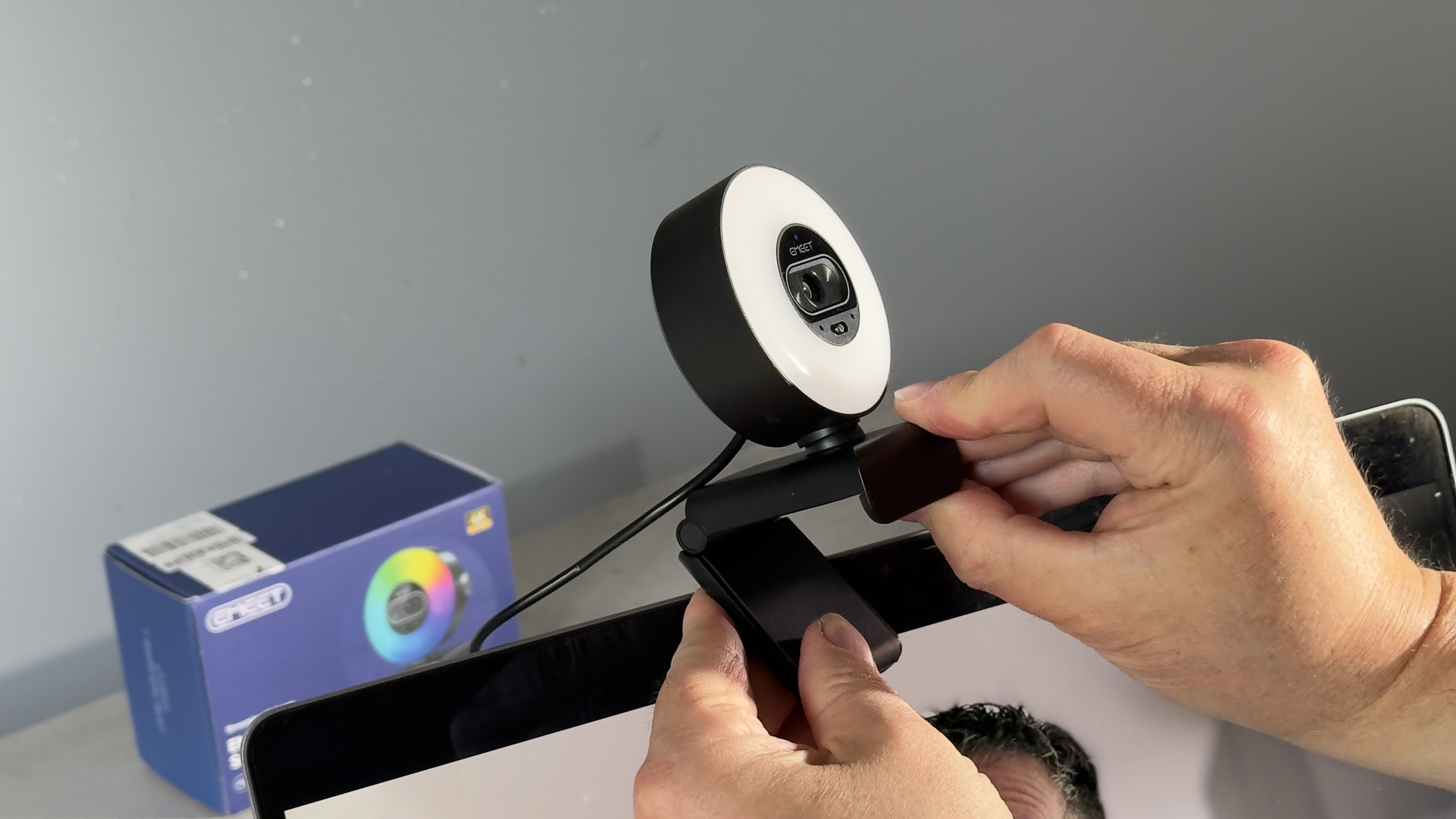 Hands attach the circular EMEET webcam and adjustable mounting clip to the top of a monitor.