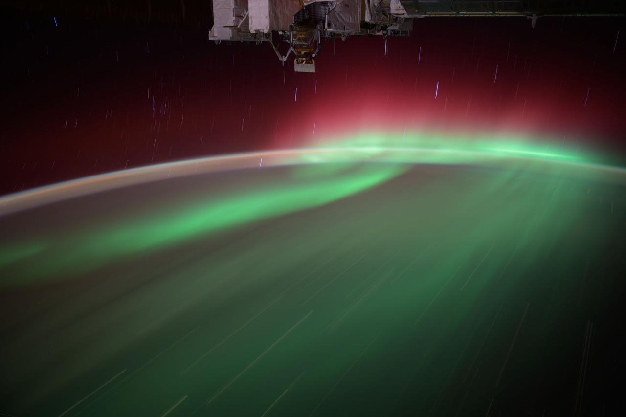 a wash of green aurora, crowned in red, wisps over the top of a night-shadowed earth.