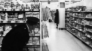 A black and white photo of a security guard crouching behind a store aisle to catch a shoplifter