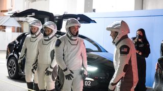 four astronauts in sleek white space suits smile and wave in front of a black SUV