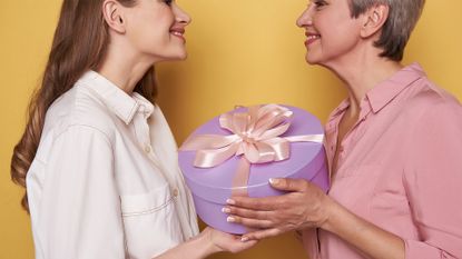 Side view of beautiful brunette girl smiling to her mummy against orange background. Old lady holding a gift in arms while looking at her daughter indoors. Mothers Day concept