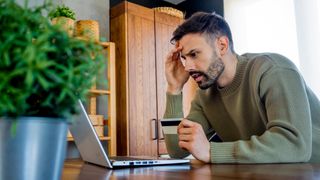  Worried man using laptop and credit card in a modern kitchen, stressed about inflation and high prices. 