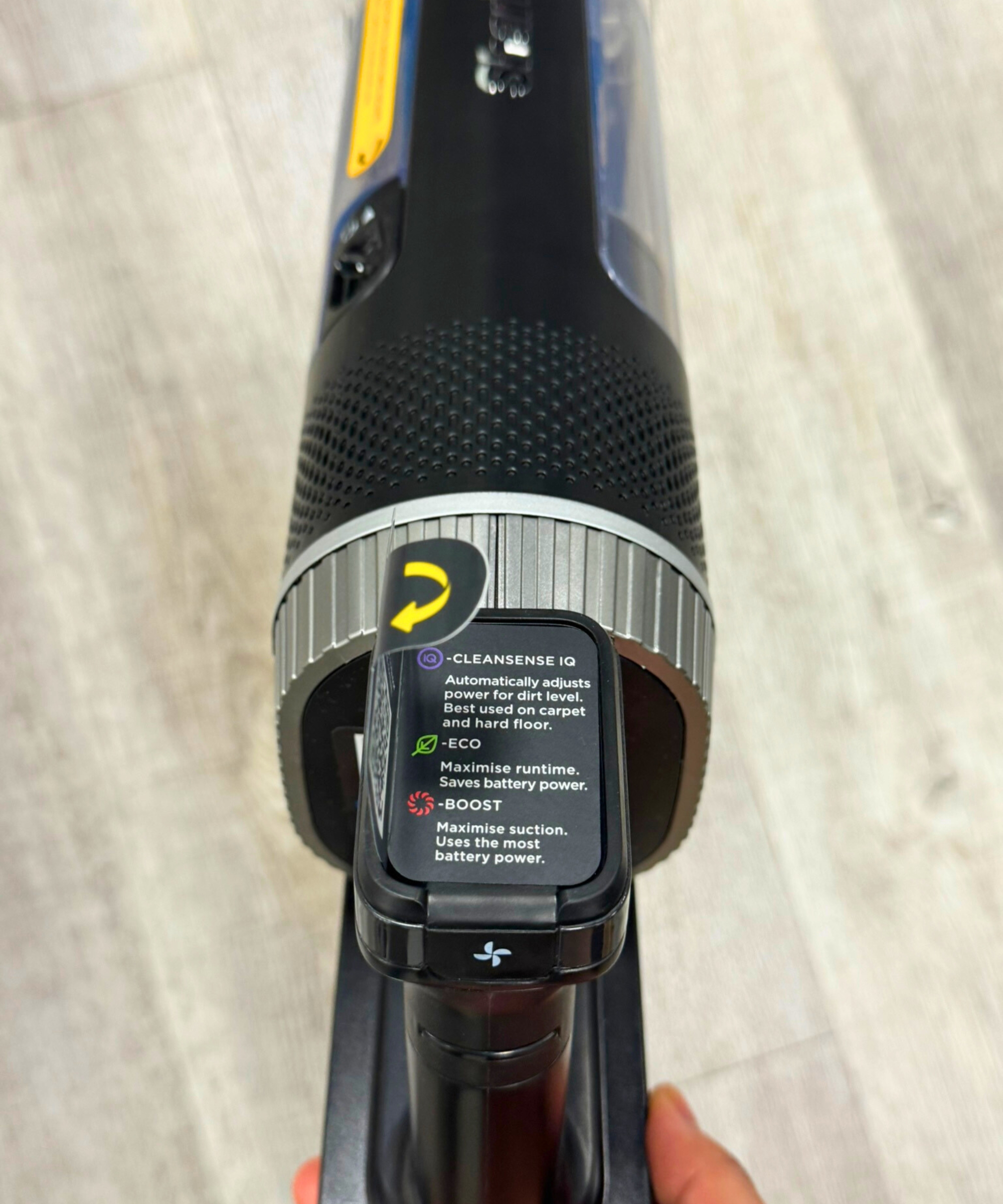 Closeup of sticker on Shark Stratos Cordless vacuum showing the suction modes available. The Shark Stratos vacuum is being held above laminate floor.
