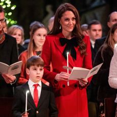 Princess Kate holding a candle wearing a red coat at church standing next to Prince Louis in a suit