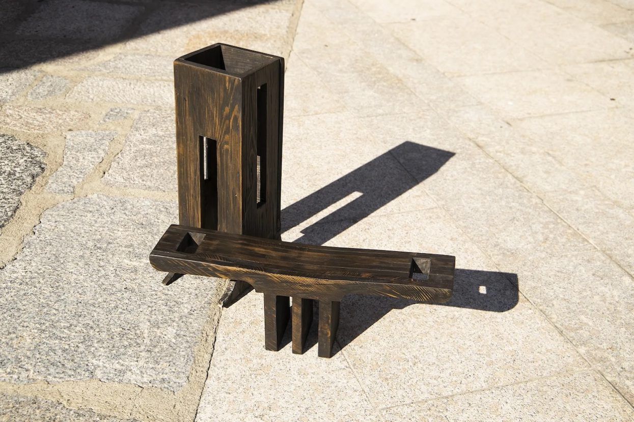 Two minimalist-cut wooden sculptures in dark tones stand out against a sun-kissed concrete pavement.