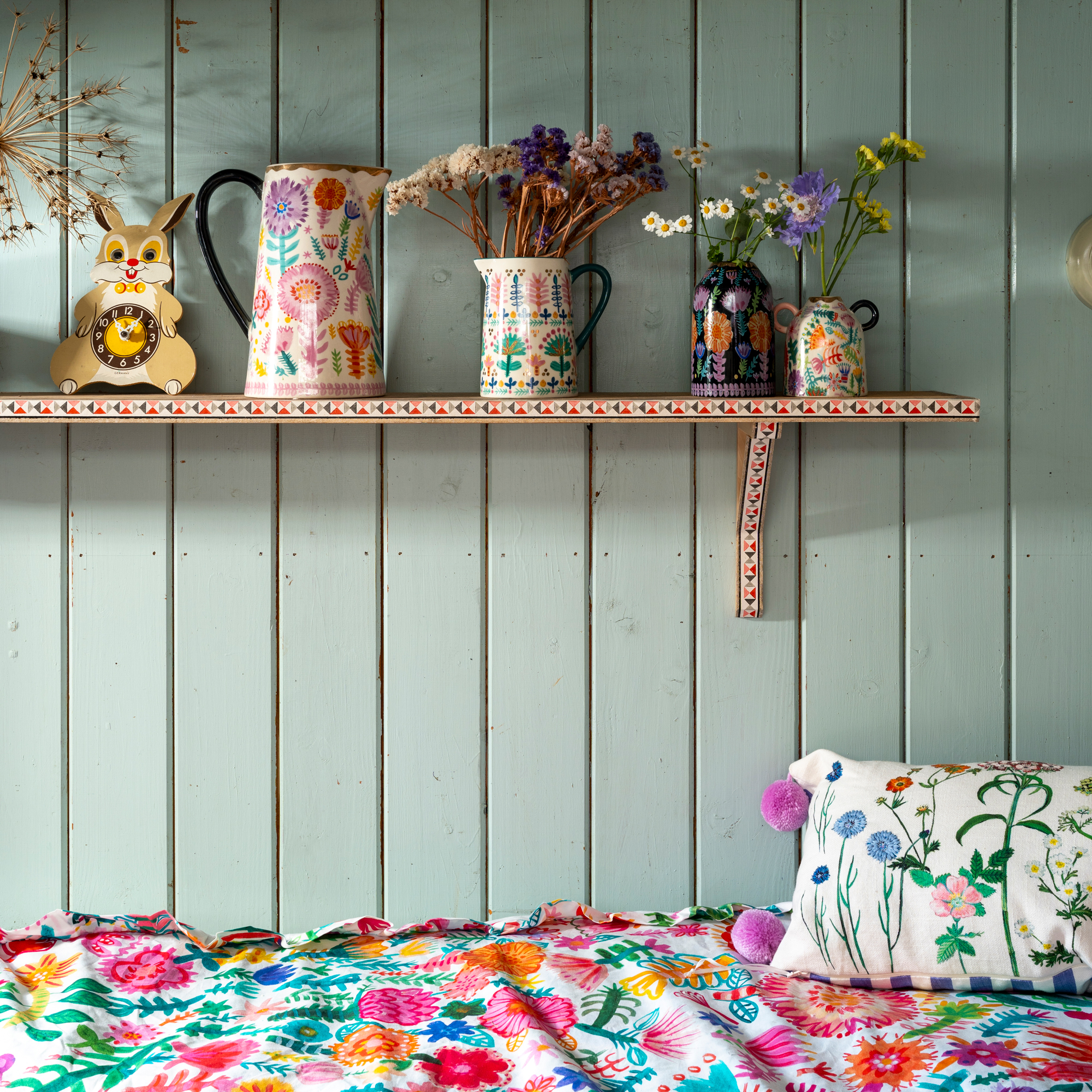 a bed beside a panelled wall with a hand-painted shelf with jugs and vases