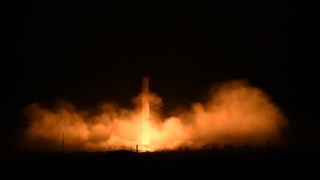 A rocket launches in the dark with an orange glowing cloud beneath it