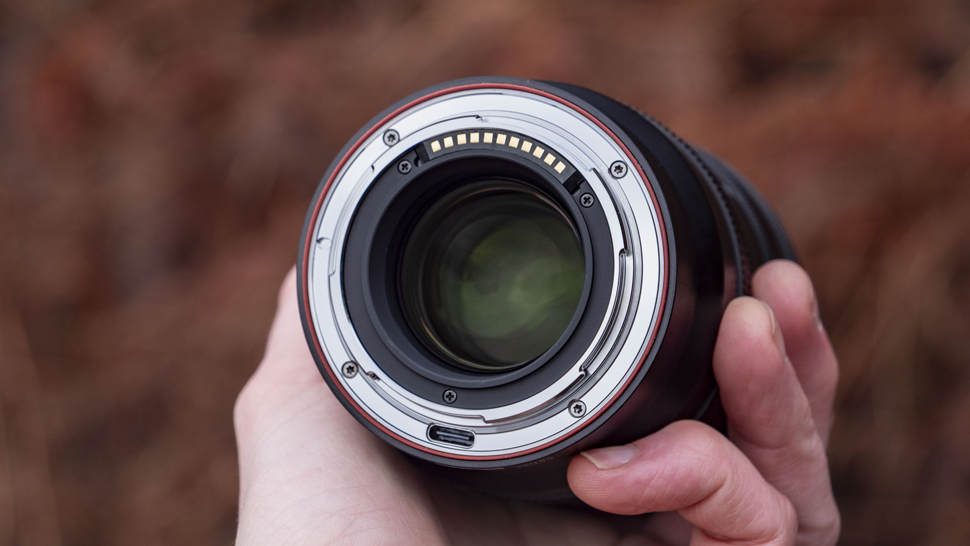 Rear mount of the Viltrox AF 85mm F1.4 Pro lens in photographer's hand, outdoors and with bronze-colored ferns in the background