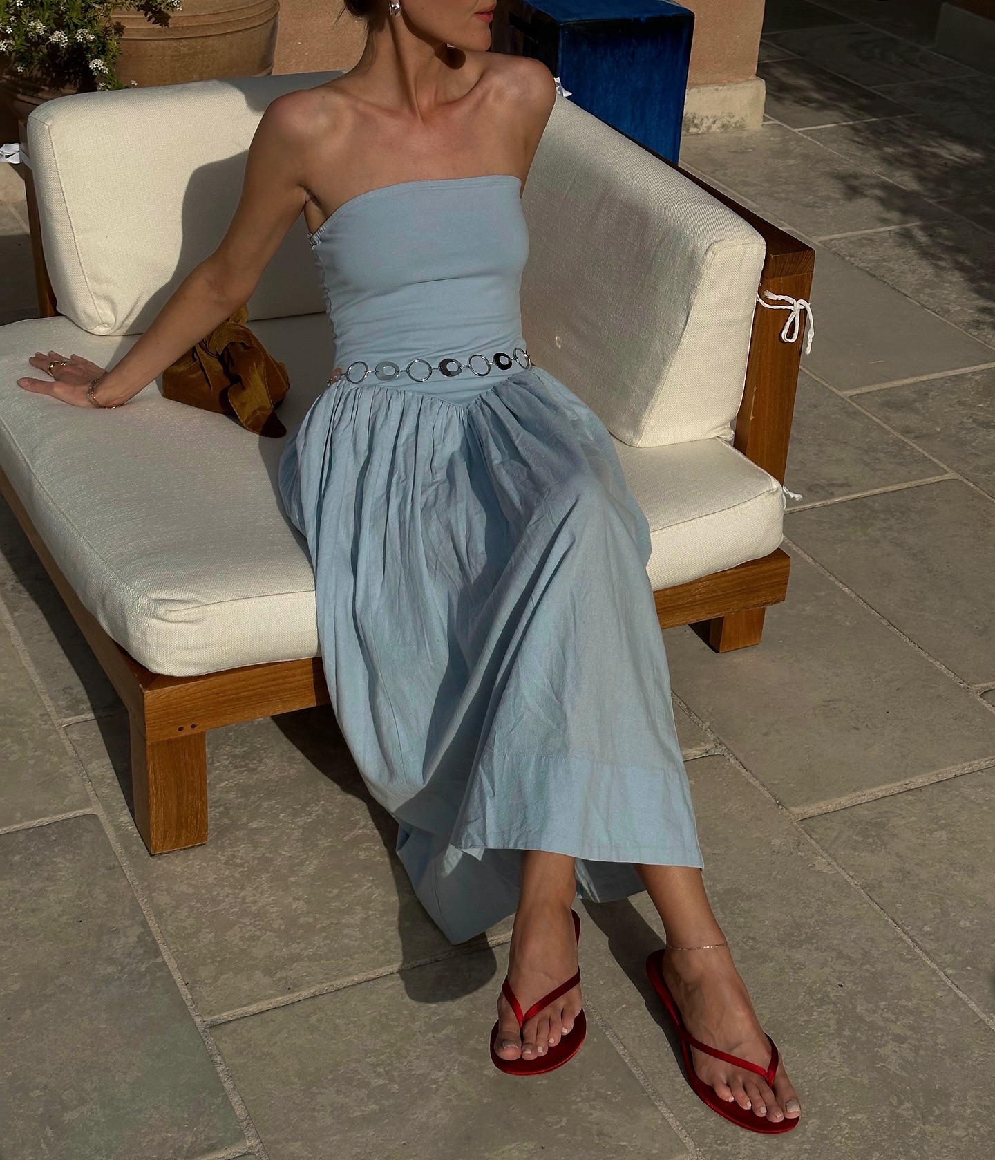 Fashion person Chloe wears a light blue dropped waist dress a chain belt and the reformation Jessie sandal in red. She is sat on a beach chair in a recent image from her Instagram.