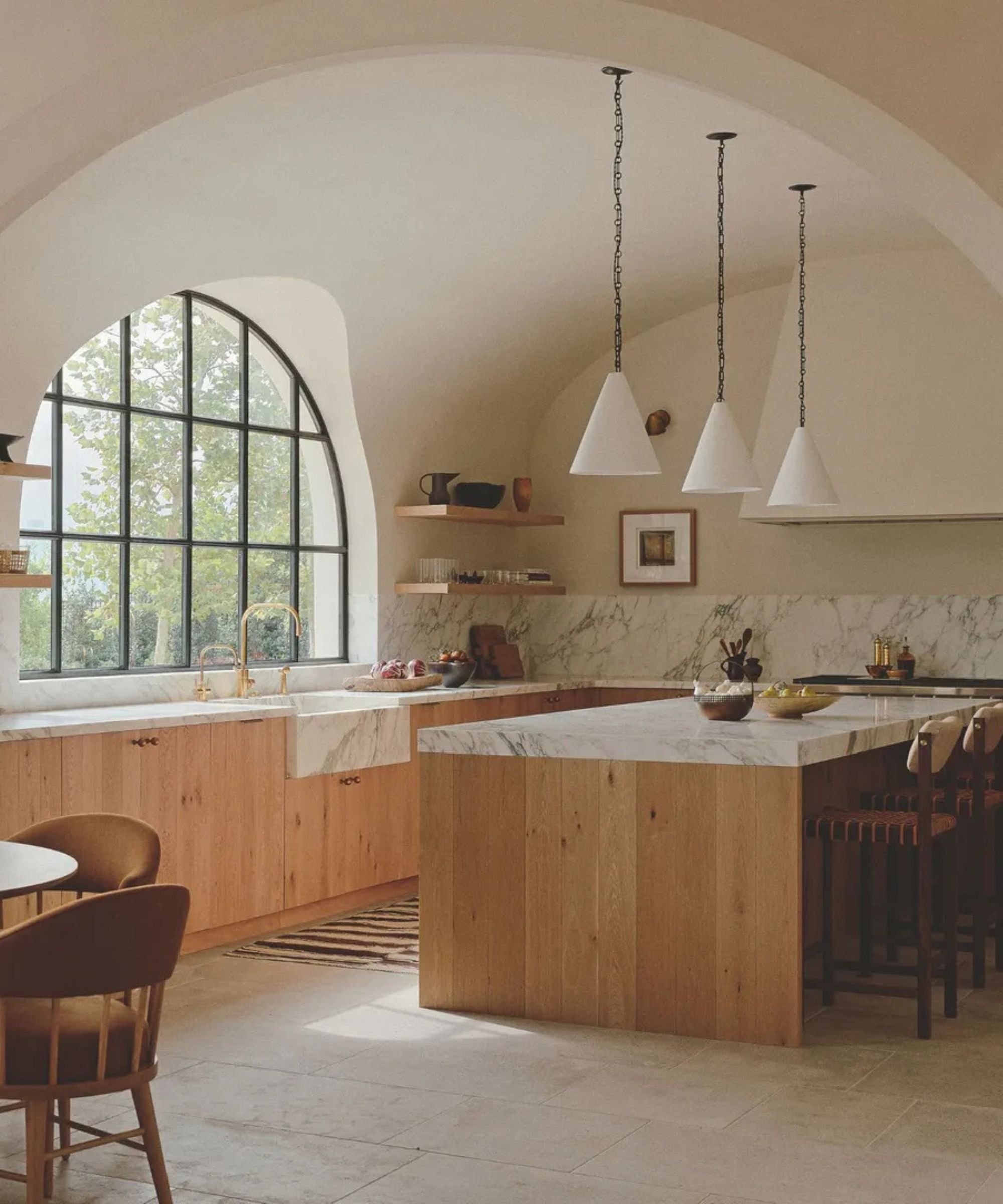 A spacious, warm-toned kitchen with a large marble-topped island, light oak cabinetry, and dramatic arched architecture. The space is illuminated by natural light from large grid windows and three white conical pendant lights