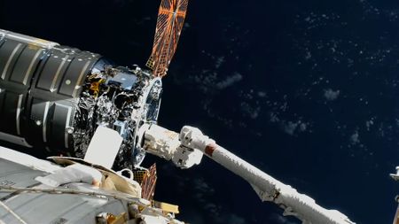 A large robotic arm reaches and grapples a silver-cylindric spacecraft with round solar arrays outside a space station.