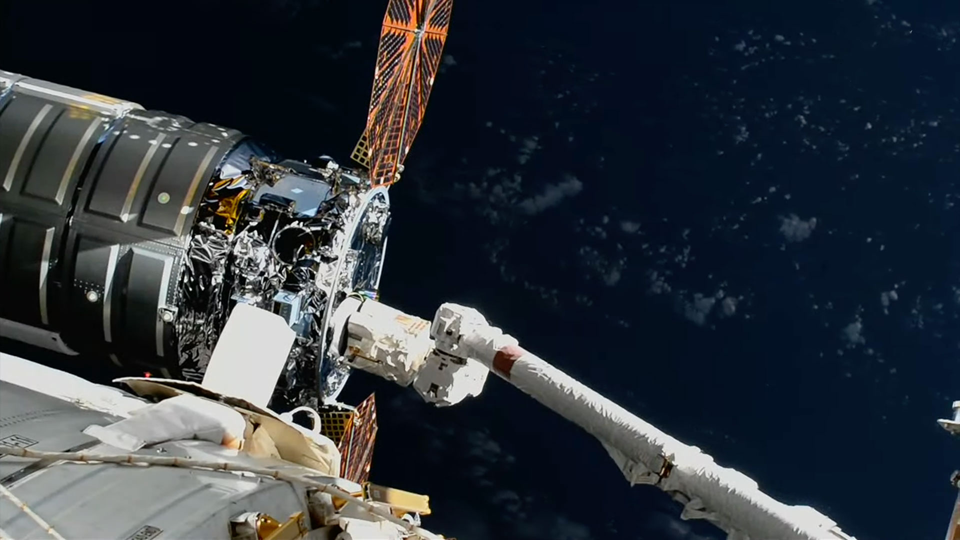 A large robotic arm reaches and grapples a silver-cylindric spacecraft with round solar arrays outside a space station.