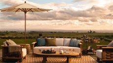 An outdoor seating area with wooden seats with white cushions, a wooden coffee table with a tea set on it, a white umbrella, and rolling hills in the background