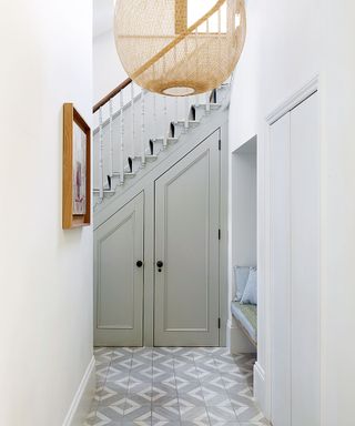 Hallway with the staircase and built in cupboards, large woven lampshade and grey and white tiled floor.