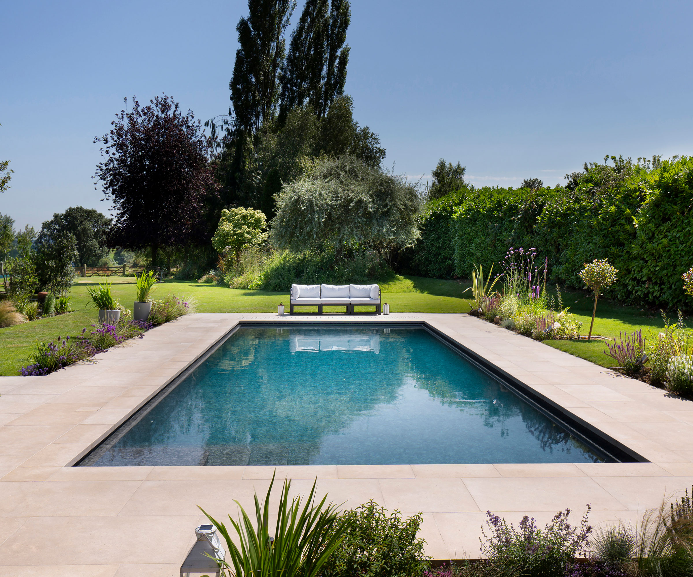 swimming pool with natural stone patio surround, garden sofa, lawn, trees, and planting, and reflections in the water