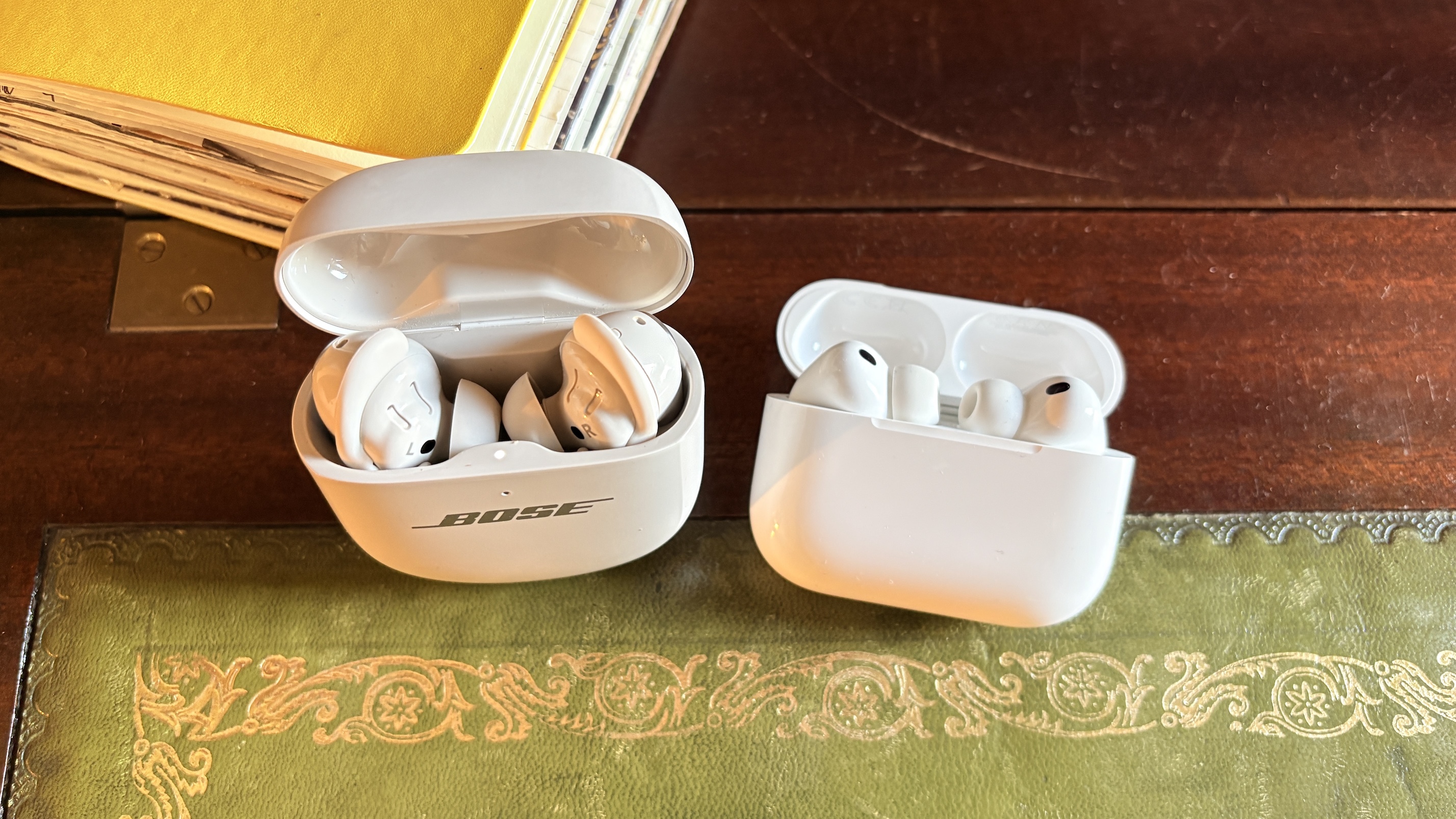 The AirPods Pro 3 and Bose QuietComfort Ultra Earbuds (2nd Gen) in their open charging cases on a green material surface part of a wooden table.