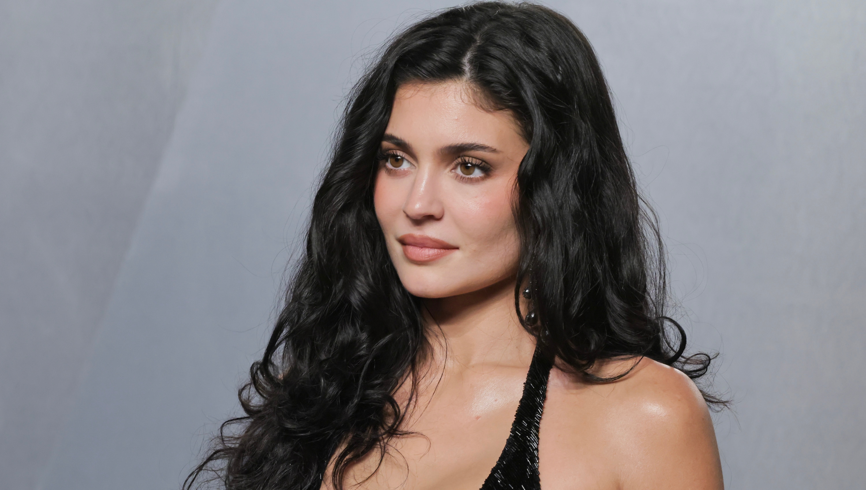 a white woman with long, dark hair poses in front of a gray background