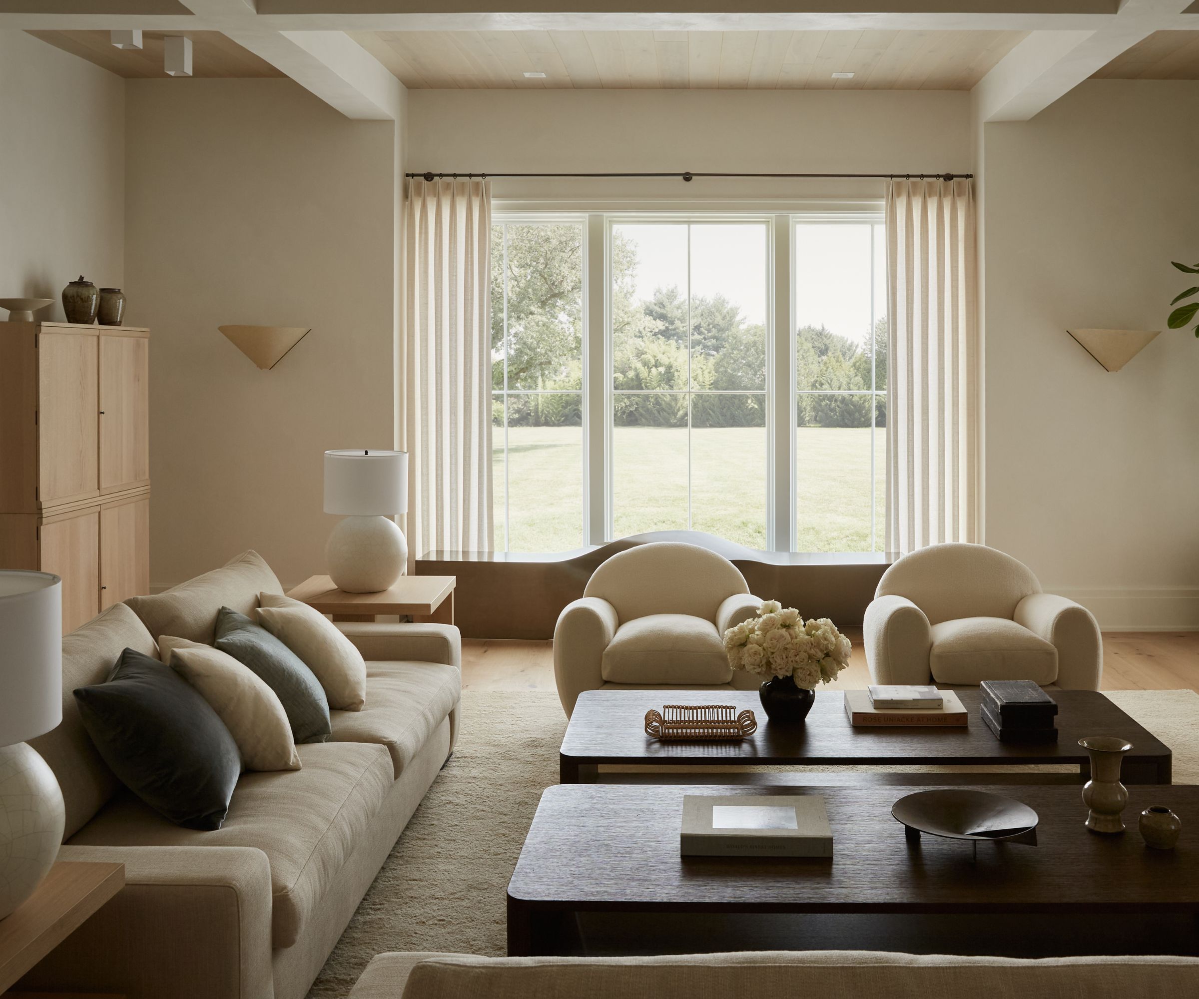 Warm neutral living room with a pair of cream velvet armchairs, two dark wood low coffee tables, a neutral velvet sofa, a large window with sheer drapes, and cream walls.