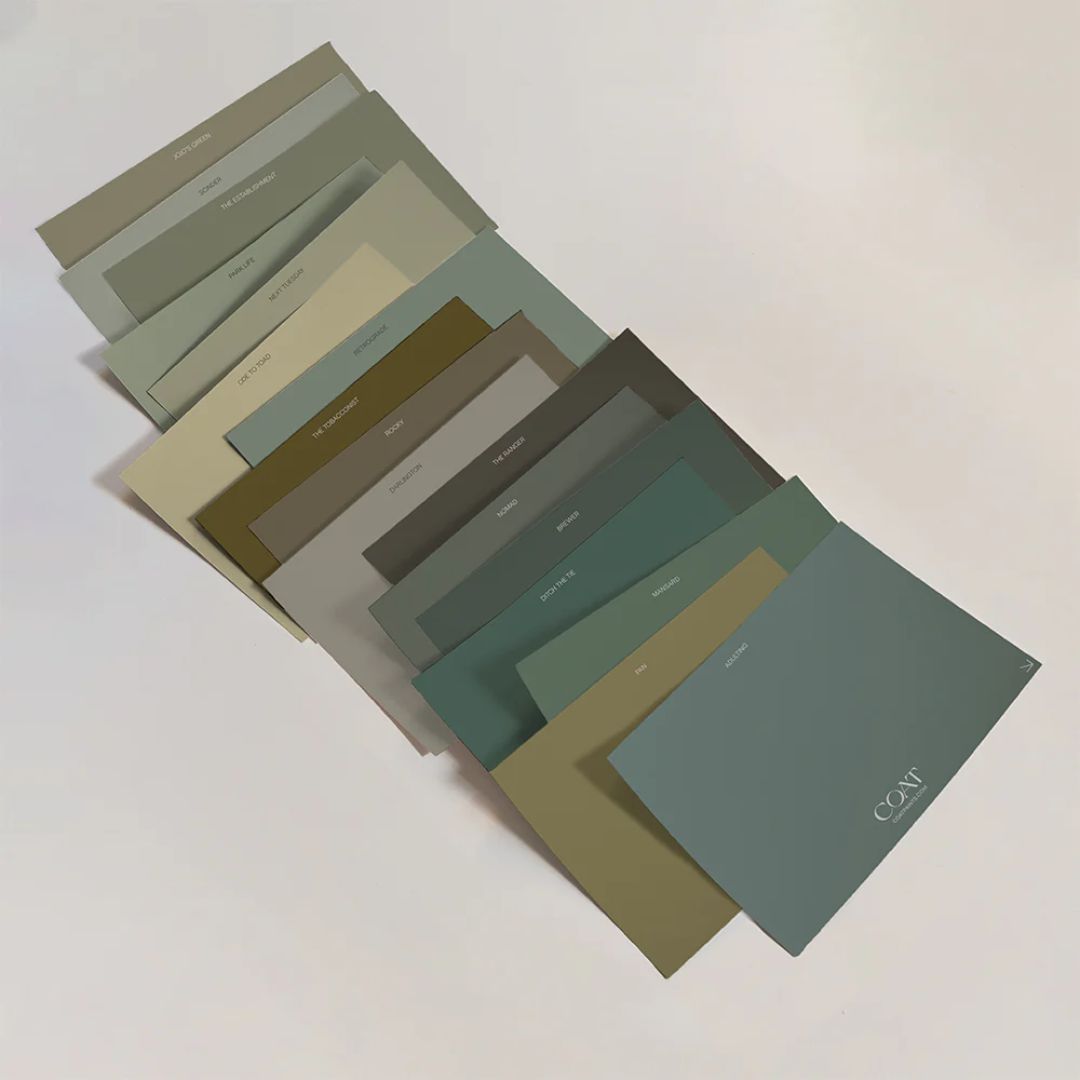 A fan of rectangular paint sample cards in muted greens, sages, olives and soft greys is spread across a white surface. Each card is labelled with shade names, and the top card shows the brand &amp;ldquo;COAT&amp;rdquo; in white lettering.