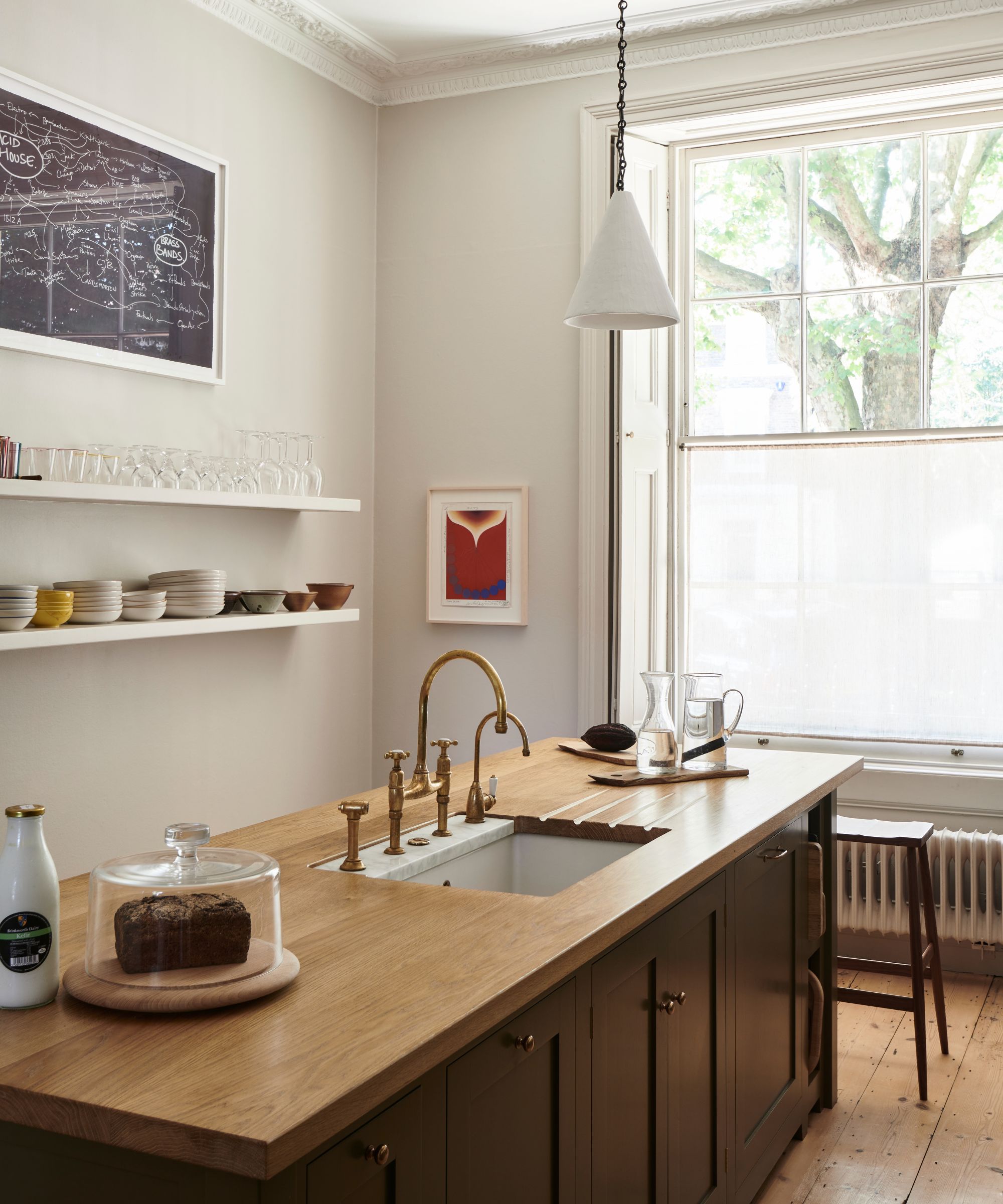Large kitchen with large window, crown moulding, a free-standing island with a wooden countertop and dark brown cabinetry with a sink and gold tap, and two open shelves with glassware and ceramics.