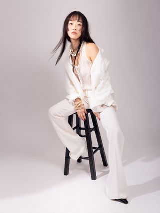 XO, Kitty actress Gia Kim poses on a black stool wearing a Mariusz Brzezinski white tank top, shirt, and wide-leg trousers with layered necklaces and bracelets.