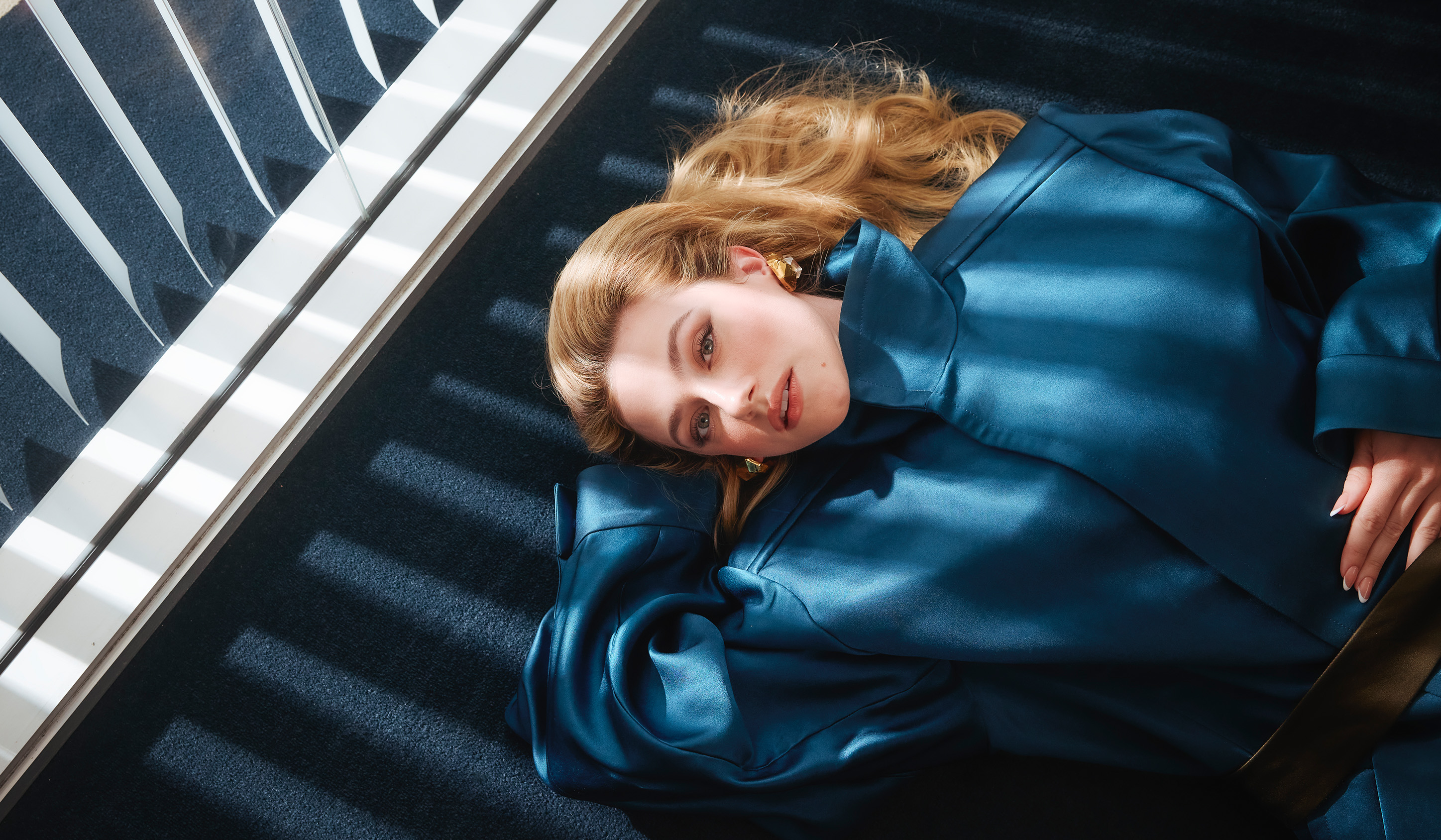 Lili Reinhart wearing a blue jacket lying on the blue carpet with one hand behind her head