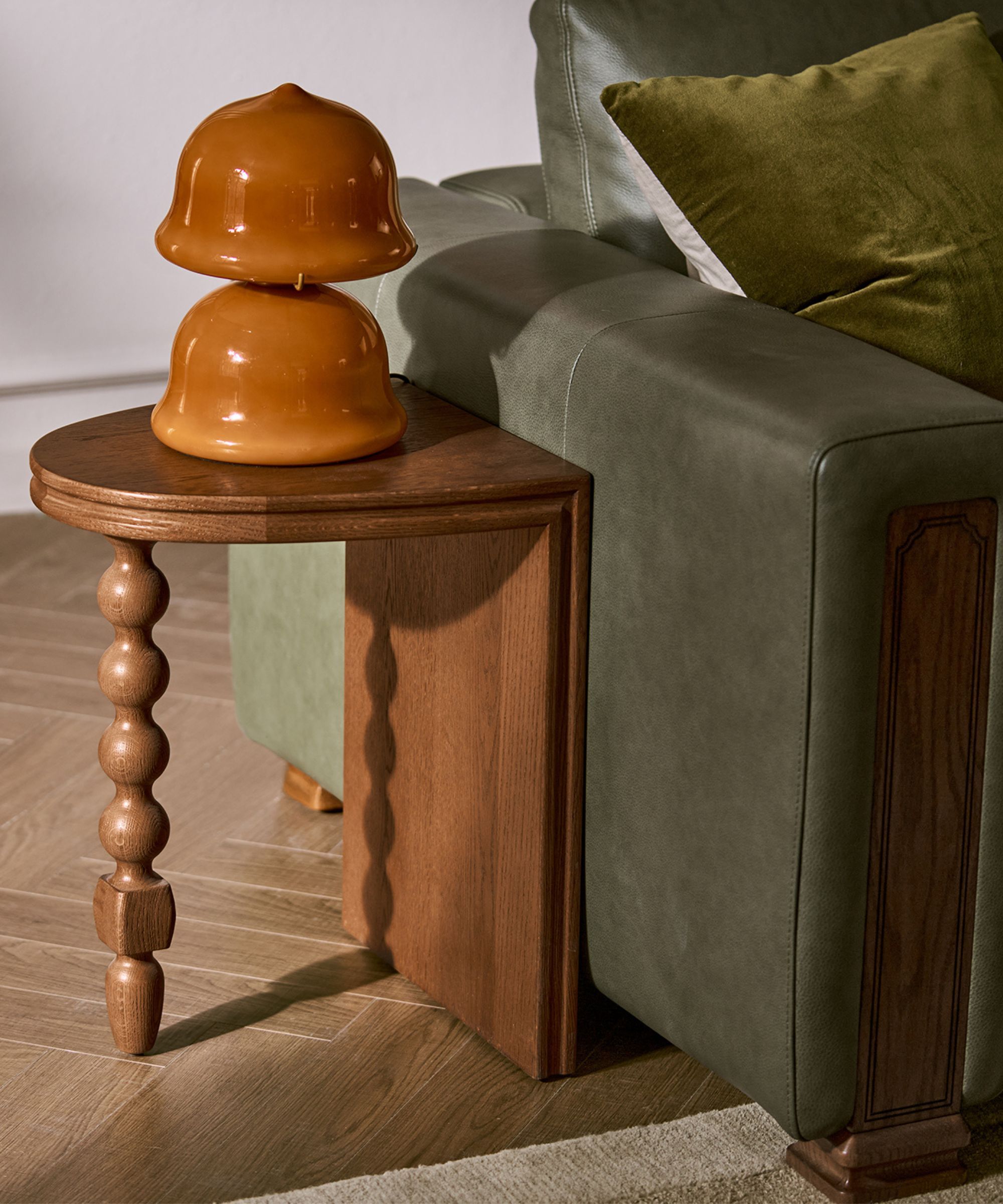 close up of a side table with one bobbin leg, an orange glass lamp, and a green sofa with timber detailing from Castlery x Tan France