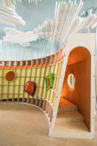 A long sweeping curved playhouse made from plywood with different coloured windows in geometric shapes.