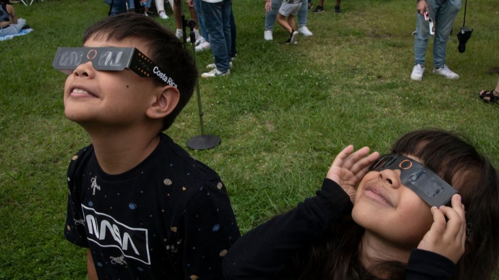 two children watching an eclipse through eclipse glasses they are both smiling.