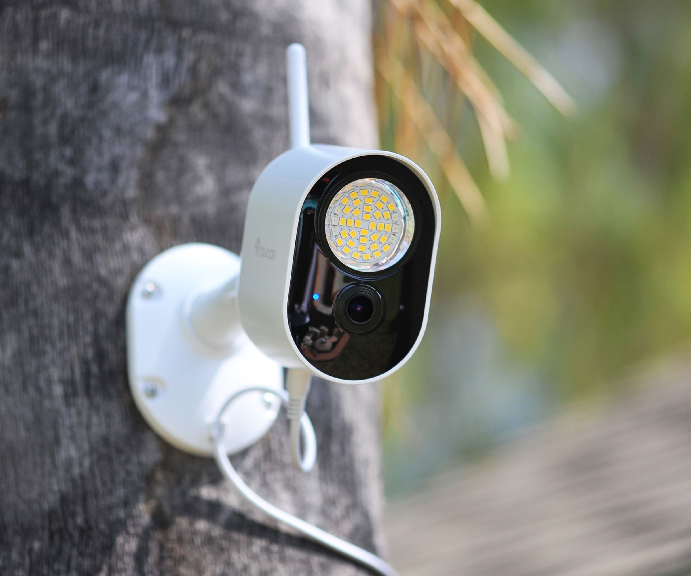 A white wireless outdoor security camera with built-in LED floodlight and Wi-Fi antenna, wall-mounted on a wooden post.