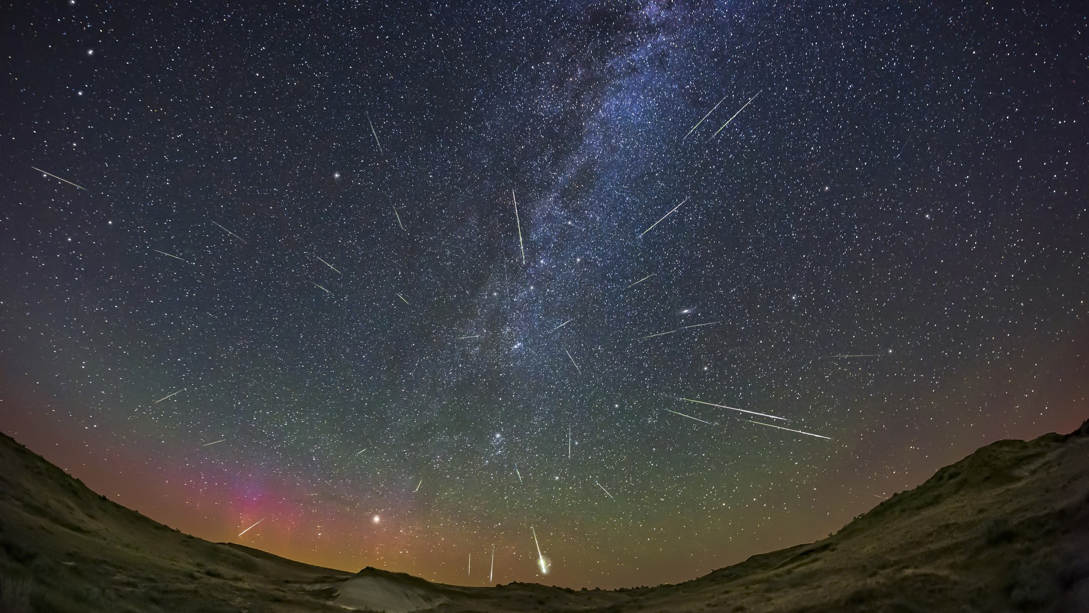 About three dozen Perseid meteors over Dinosaur Provincial Park, Alberta, Canada. A dim magenta aurora is visible to the northeast at left. Cassiopeia is at centre above the radiant point; the Andromeda Galaxy is just right of centre. Capella is rising at left. Airglow also tints the sky.