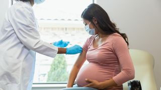 a pregnant woman wearing a pink shirt and blue surgical mask sits as a standing medical provider prepares to give her a vaccine in her right arm