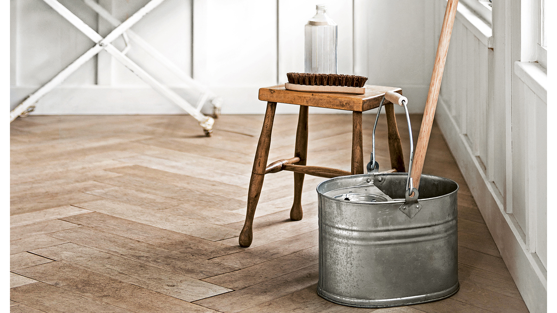 Room with wooden flooring steel bucket with a mop next to a wooden stool with cleaning solution on it