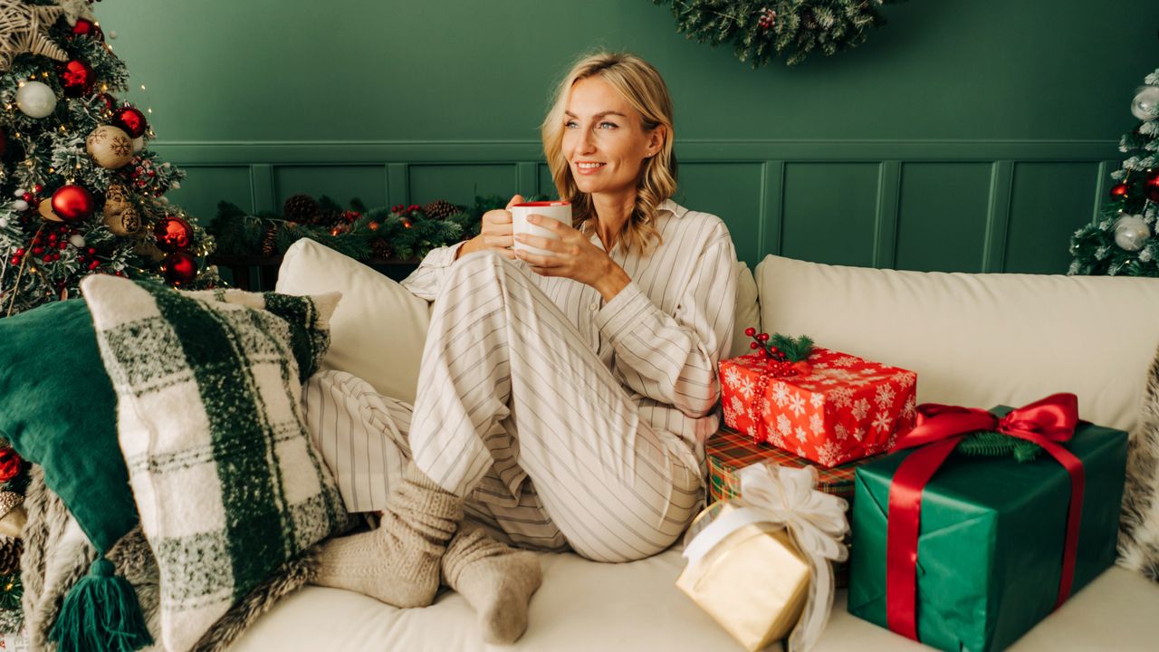 A beautiful blonde woman is sitting on the sofa in the Christmas living room and drinking coffee. - stock photo