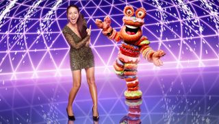 Davina McCall stands in front of a purple background next to Masked Singer character Doughnuts — a costume made up of rings of doughnuts stacked on top of each other
