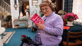 Mrs Brown's Boys 2025 publicity image. Brendan O'Carroll as Mrs Brown reading a book and on a mobility scooter. 