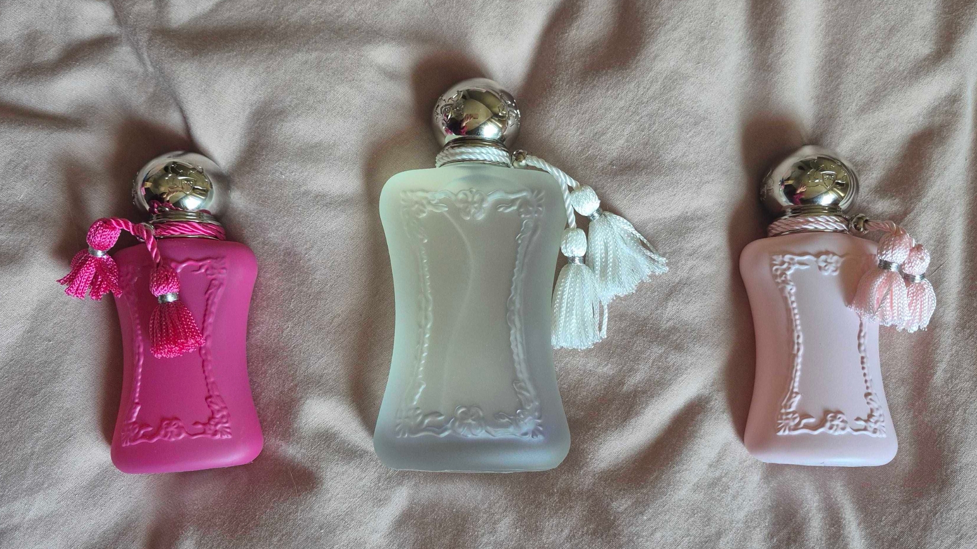 A picture showing some of the Parfums de Marly scents featured in this round-up, including 30ml bottles of Delina and Oriana and a 75ml bottle of Valaya, spread across a pink bedding background.