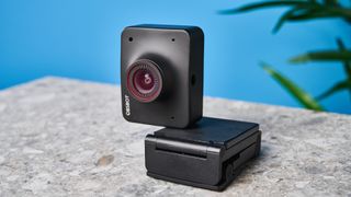 a black small 4K webcam with a red button photographed on a beige surface against a blue background