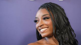 Simon Biles smiling off to one side in front of a purple backdrop