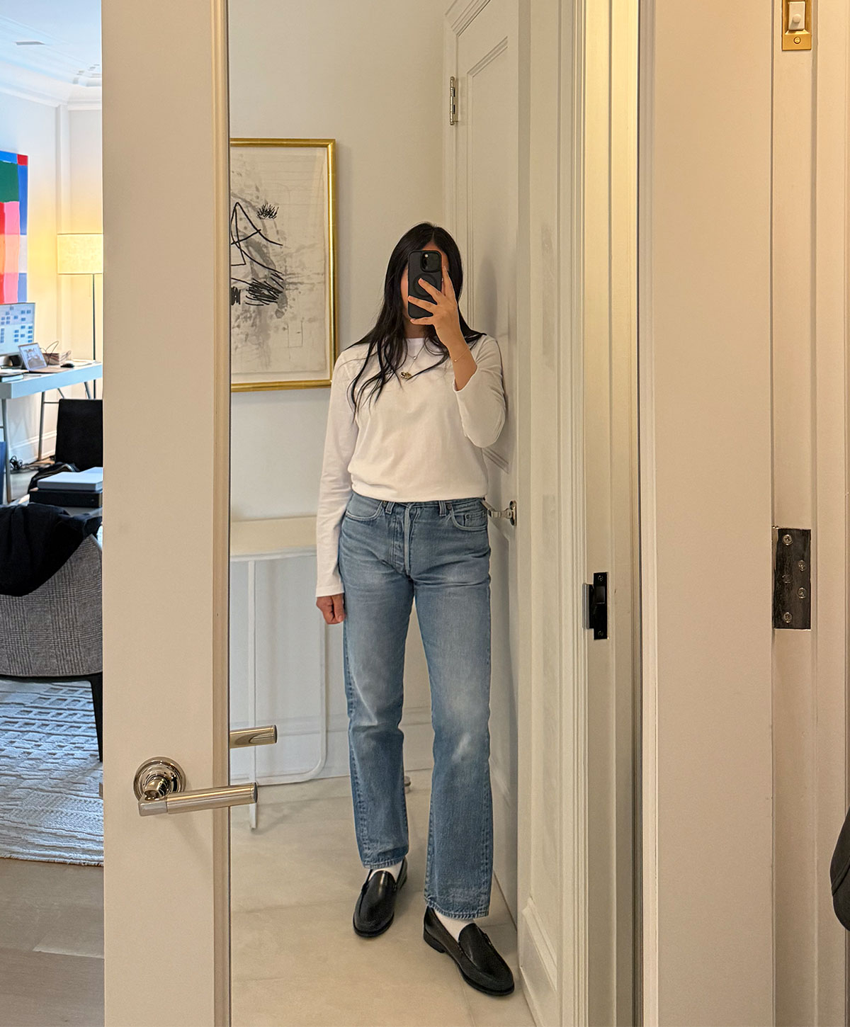 @nicoleakhtarzad taking a mirror outfit selfie and wearing a white long sleeve tee with blue jeans and loafers.