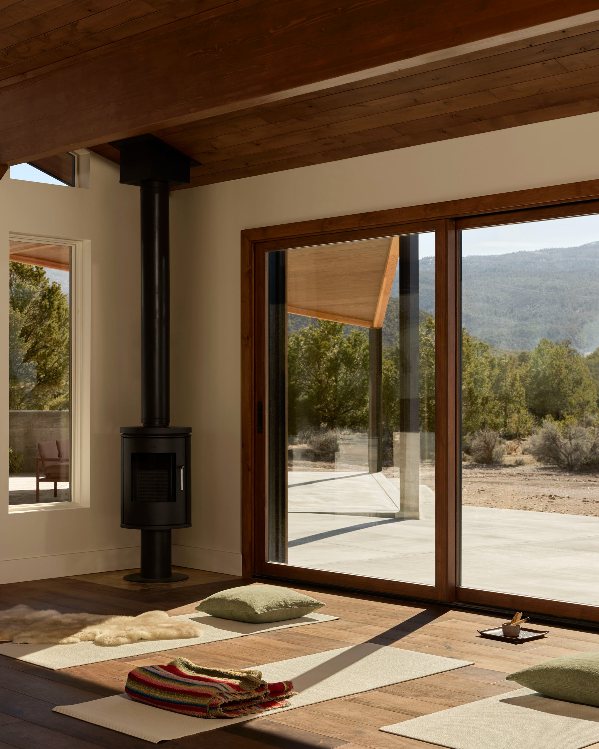 A modern private yoga studio with three mats on the floor with throw blankets in front of a closed sliding door with sweeping views of the desert vista with a black wood burner in the corner