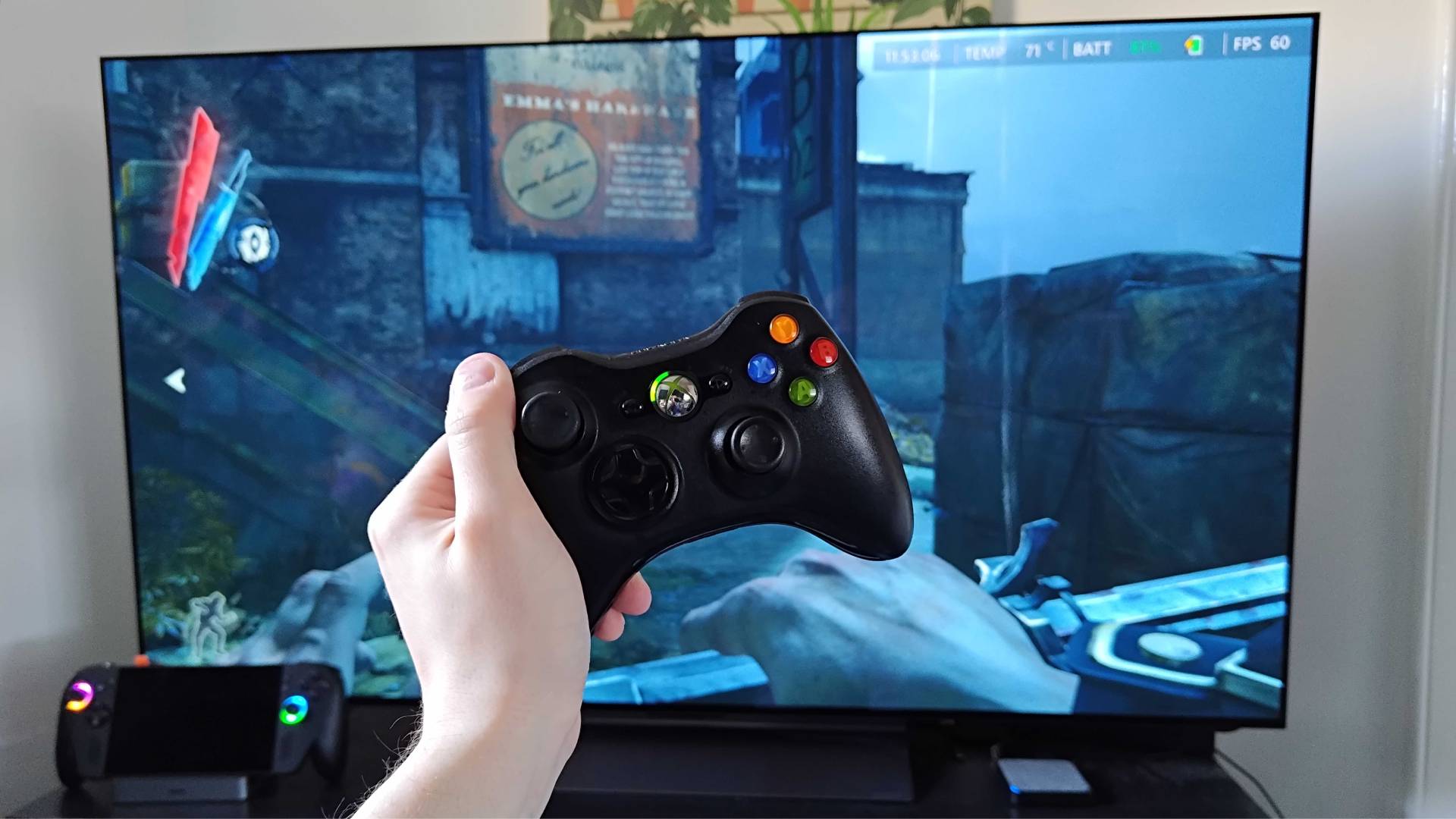 Hand holding black Xbox 360 controller in front of ROG Xbox Ally X connected to LG OLED TV with Dishonored gameplay on screen.