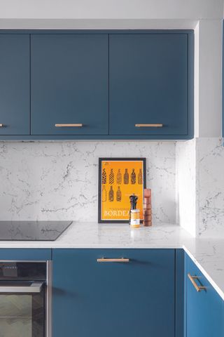 A small kitchen area with bright blue cabinets with slim gold coloured handles, and marbled tiled walls and worktops