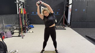 Woman demonstrates overhead side bend exercise in gym. She holds a kettlebell in both hands above her head and is leaning to one side