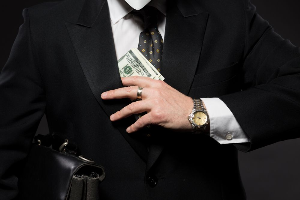 Businessman slipping money beneath suit jacket on dark gray background. 