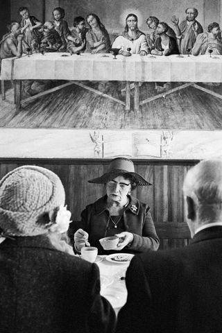 Three older people having tea at a table beneath a painting of the Last Supper