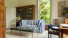 A large sitting room with a blue and white striped sofa, a floral fabric coffee table and in the background a large chest filled with ceramics. 